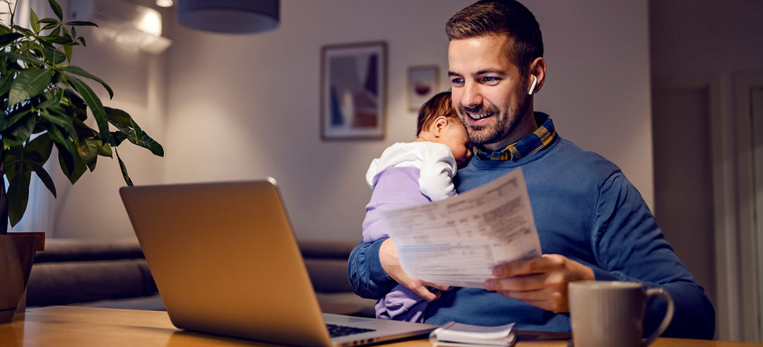 Hombre trabajando con el ordenador, sosteniendo en un brazo a un bebé y en el otro un documento